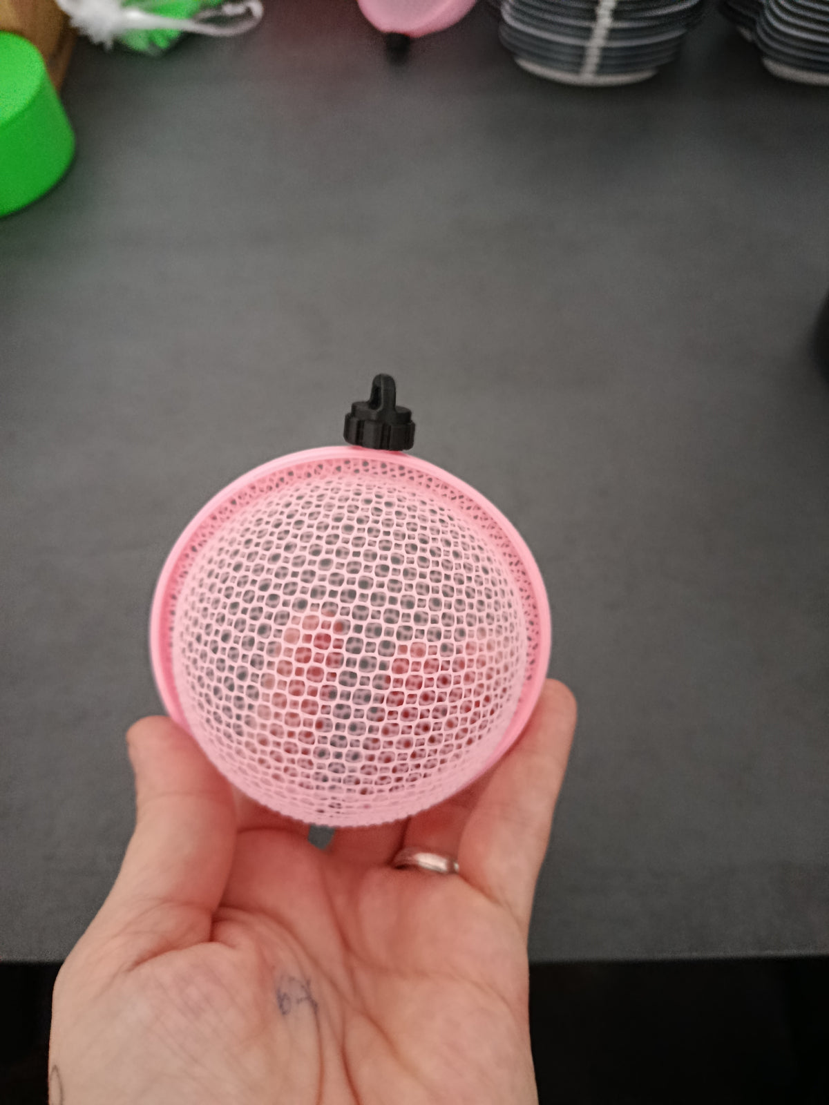 vor grauem Hintergrund: Rosa 3D-gedruckte Weihnachtskugel mit feinem Gittermuster und schwarzem Aufhänger auf einer Hand vor grauem Hintergrund.
