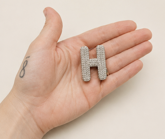 ilberfarbener Buchstaben-Magnet H in Strickoptik in einer Hand als Größenvergleich